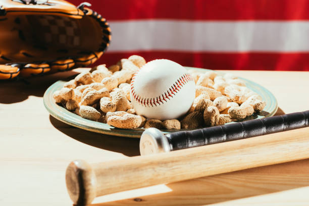 zbliżenie kijów baseballowych, piłka baseballowa na talerzu z orzeszkami ziemnymi i skórzaną rękawiczką na drewnianym stole z flagą usa za - baseball baseballs peanut american culture zdjęcia i obrazy z banku zdjęć