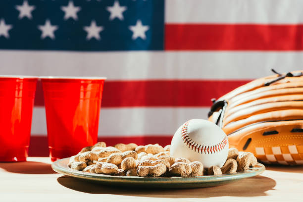 zbliżenie piłki baseballowej na talerzu z orzeszkami ziemnymi, czerwonymi plastikowymi kubkami i rękawicą baseballową na stole z flagą usa za - baseball baseballs peanut american culture zdjęcia i obrazy z banku zdjęć