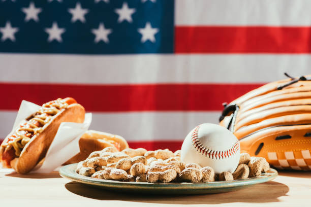 zbliżenie piłki baseballowej na talerzu z orzeszkami ziemnymi, skórzanymi rękawiczkami i hot dogami na stole z flagą usa za - baseball baseballs peanut american culture zdjęcia i obrazy z banku zdjęć