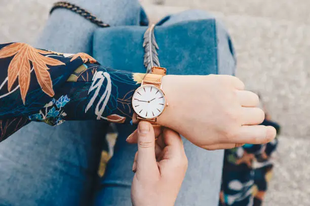 Photo of close up, young fashion blogger wearing a floral jacker, and a white and golden analog wrist watch. checking the time, holding a beautiful suede leather purse.