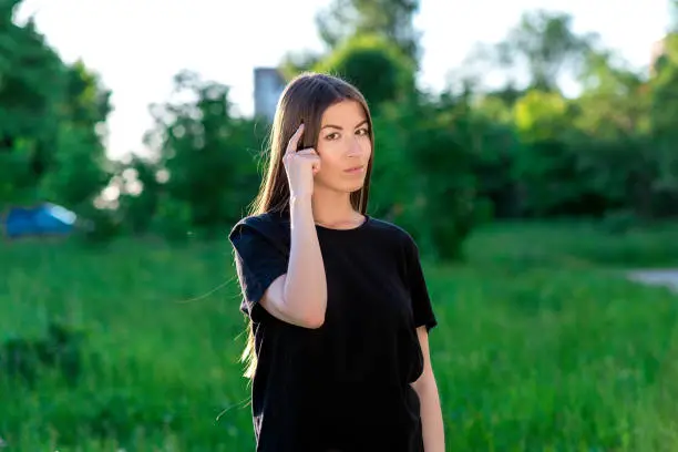 Photo of The brunette girl. Summer in the park in the fresh air. Pointing to your head with your finger indicates. Emotionally shows think. Right thoughts and Fantasies.