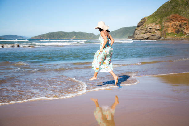 Beautiful woman jumping a small wave of the sea, happily, in a flowery dress and hat, on a sunny day. stock photo
