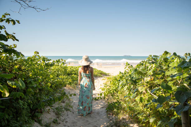Beautiful woman walks on a path lined with vegetation towards the sea, on a deserted beach, on a sunny day. stock photo