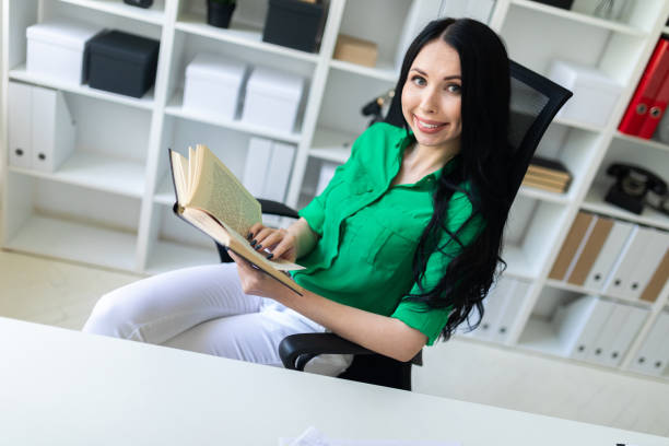 A young girl is sitting at the office desk and is holding a book. Beautiful girl in green blouse and white pants running in the office. photo with depth of field. benefits of reading book stock pictures, royalty-free photos & images