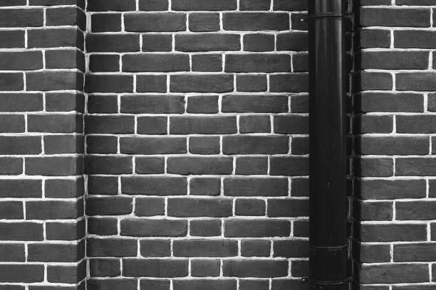 istock Black brick wall with black downspout. Drain pipe is in right. Bright geometric and symmetrical background image of monochrome brick wall. 972349800