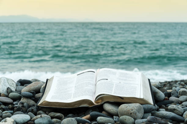 Bible open in front of the sea. Bible open on top of small stones in front of the green sea and an orange sky. Copy space. new testament stock pictures, royalty-free photos & images
