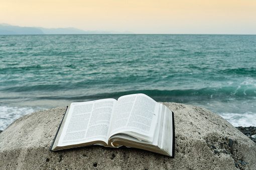 Open bible on top of a rock in front of the sea of greenish waters and an orange sky. Copy the space.