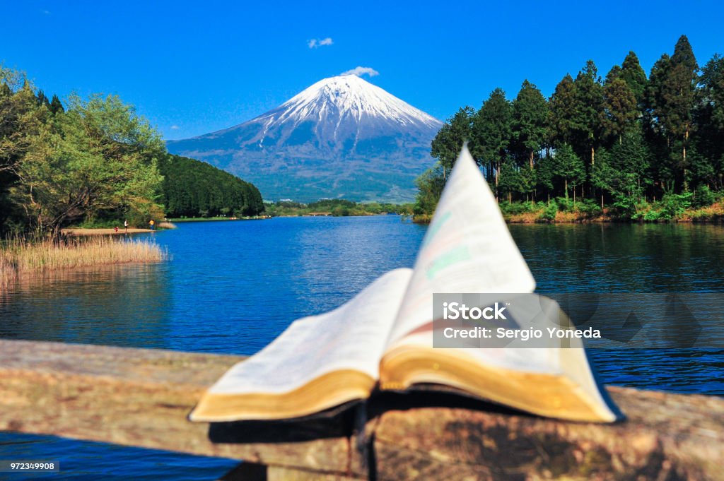 Bible open in front of the majestic Mount Fuji. Open bible on top of a wooden parapet in front of the beautiful Mount Fuji. A beautiful blue sky day. Pages fluttering in the wind. Tanuki lake, Japan. Bible Stock Photo