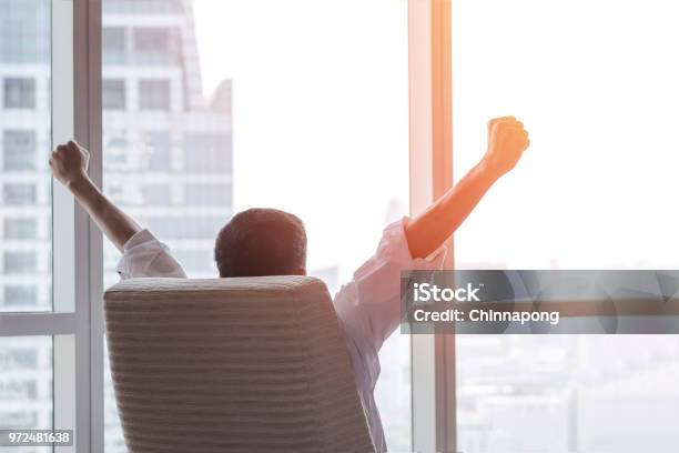 Business Achievement Concept With Happy Businessman Relaxing In Office Room Resting And Raising Fists With Ambition Looking Forward To City Building Urban Scene Through Glass Window Stock Photo - Download Image Now