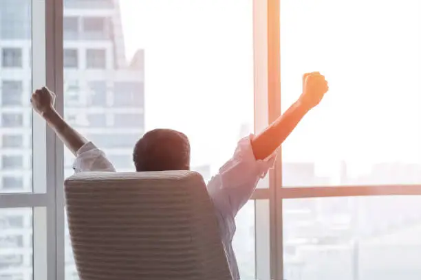 Photo of Business achievement concept with happy businessman relaxing in office room, resting and raising fists with ambition looking forward to city building urban scene through glass window