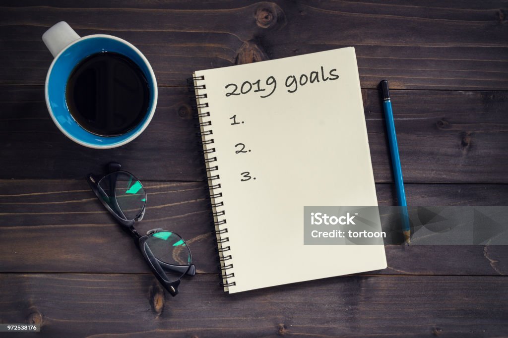 Notebook con metas 2019 masaje, lápiz, vasos y taza de café sobre fondo de madera. - Foto de stock de Aspiraciones libre de derechos