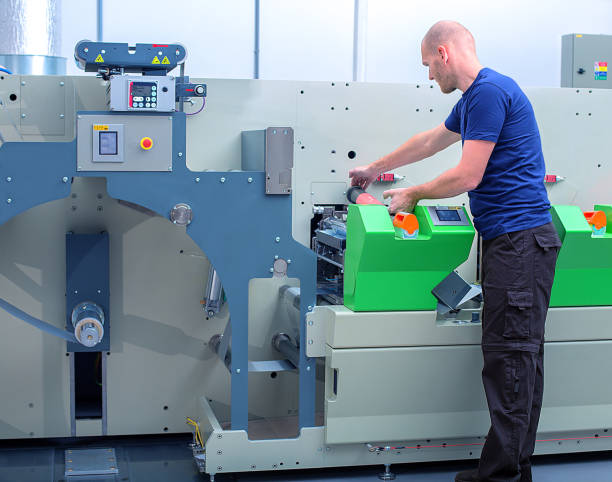 istock Printer operator next to the printing machine, holding printing cylinder with polymer relief plate stuck on it. Scene showing the preparation for the printing process on in-line press machine. 972553720