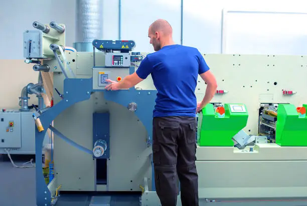 Photo of Worker next to the printing machine inputs the data by pressing the touch screen. Skilled printing operator controls printing machine via the touch screen. Rotary printing press. In-line press machine.