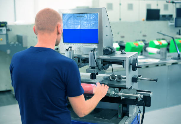 istock Worker in a printing and press center uses plate mounting machine to attach polymer relief plate on a printing cylinder. Scene showing mounting of printing plate for flexographic print machine. 972557190