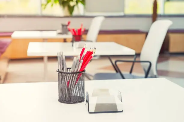 Close up desk with focused stand with stationery, pens and pencils with blurred background of modern light empty open space office. Coworking workplace concept. Minimalism business style. Copy space