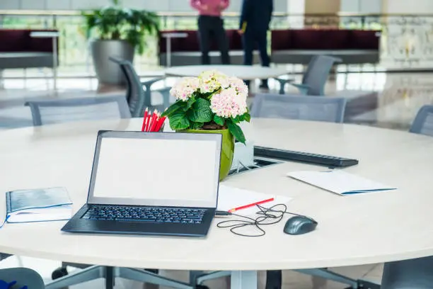 Modern stylish work place with laptop with blank white screen, notebook, stationery and flower pot on the desk at open space office. Minimalism business style. Coworking. Copy space. Selective focus