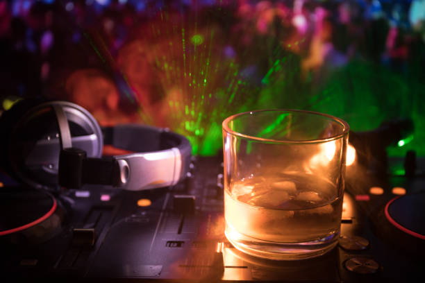 istock Glass with whisky with ice cube inside on dj controller at nightclub. Dj Console with club drink at music party in nightclub with disco lights. 972572522