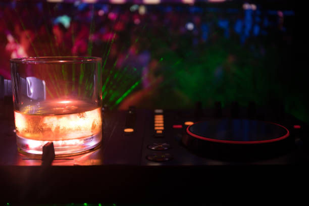 istock Glass with whisky with ice cube inside on dj controller at nightclub. Dj Console with club drink at music party in nightclub with disco lights. 972572534