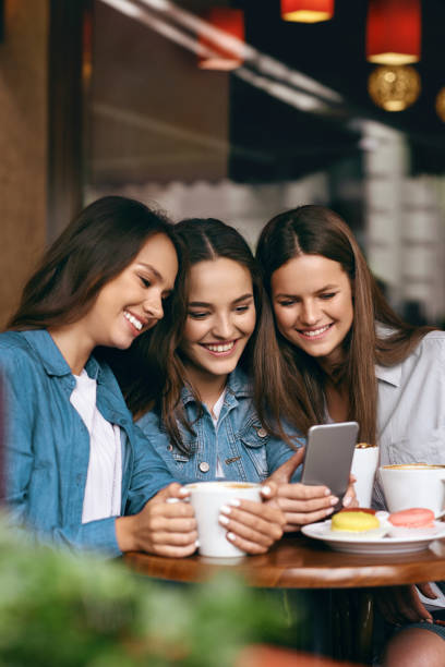 istock Young Female Friends Using Phone And Drinking Coffee In Cafe. 972655420