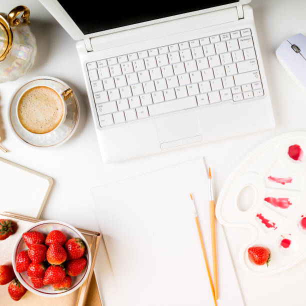 Flat lay trendy feminine home art workspace. Home office desk with laptop, notebook, paints, brushes, canvas, fresh strawberries and coffee cup on white background