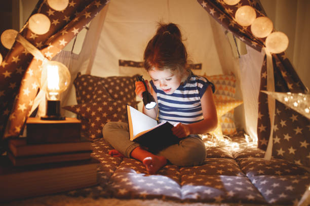 szczęśliwa dziewczynka śmieje się i czyta książkę w ciemności w namiocie w domu - child bedtime imagination dark zdjęcia i obrazy z banku zdjęć