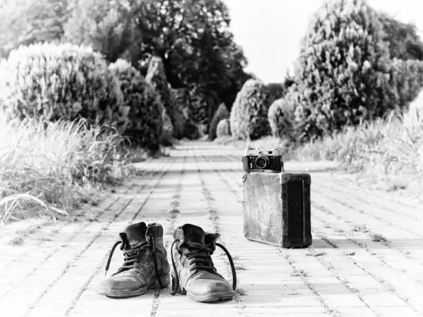 Old ankle leather boots in a foreground, in the middle of a brick road. A film camera on an antique cardboard suitcase with corner protectors, in a blurry background. A black-and-white photo effect.