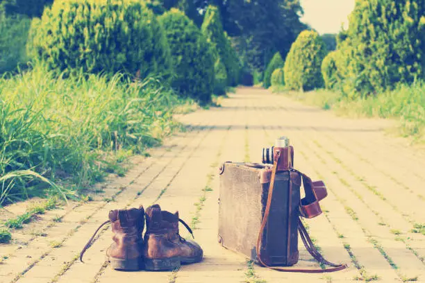 Shabby ankle boots next to a vintage cardboard suitcase, a film camera in its open leather case, and a yellow brick road, grass, and trees in a blurry background. Post-processed.