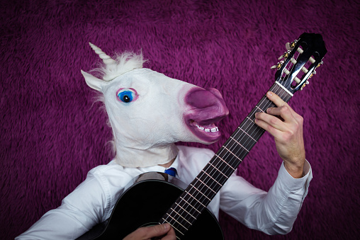 Freaky young man in comical mask playing the guitar on the purple background. Portrait of unusual guy in shirt and tie with musical instrument. Musical performance.