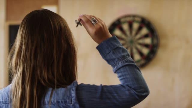 A Young Caucasian Woman with a Ring and Long Brown Hair Aims and Throws Darts at a Dartboard