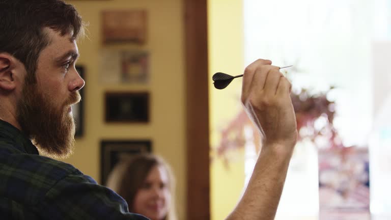 A Twenty-Something Caucasian Man with a Beard Aims and Throws Darts as a Young Brunette Woman Watches in the Background at a Bar