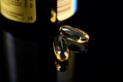 An open bottle of fish oil with the dietary supplement capsules in front of the bottle shot in studio against a black background on a reflective surface.