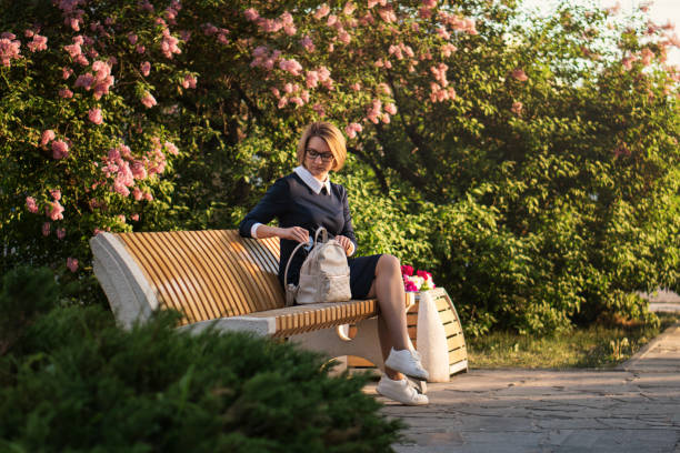Attractive girl is sitting on park bench. She is looking for something in her backpack.