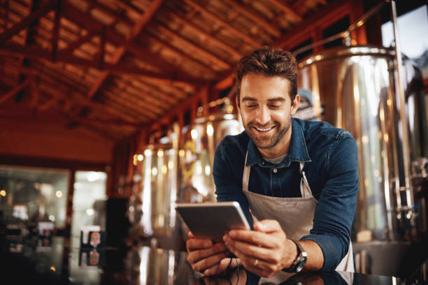 Buying time while waiting for customers Shot of a cheerful young barman browsing on a digital tablet while patiently waiting at the bar for customers inside of a beer brewery during the day artisanal food and drink photos stock pictures, royalty-free photos & images