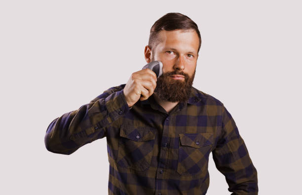 istock Making his beard perfect. Confident young bearded man shaving with electric razor and looking at camera in checkered shirt while standing against grey background 973116564