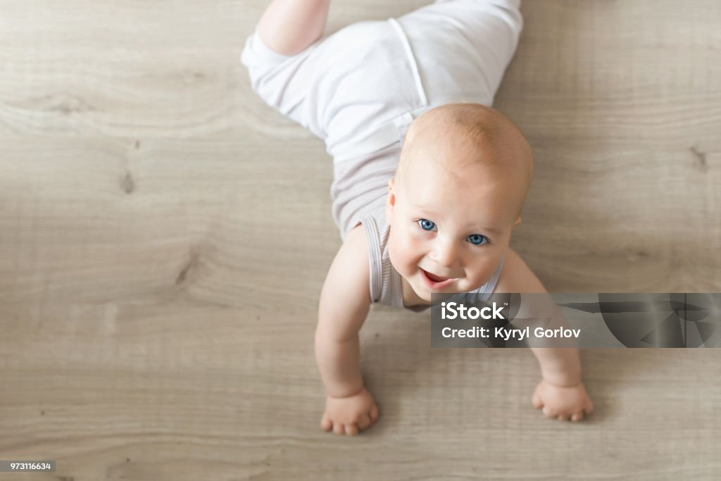 Lindo pequeño bebé acostado en madera dura y sonriente. Niño arrastrándose sobre el suelo de madera parqué y mirando hacia arriba con cara feliz. Vista desde arriba. Copyspace - Foto de stock de Bebé libre de derechos
