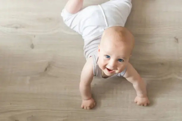 Cute little baby boy lying on hardwood and smiling. Child crawling over wooden parquet and looking up with happy face. View from above. Copyspace.