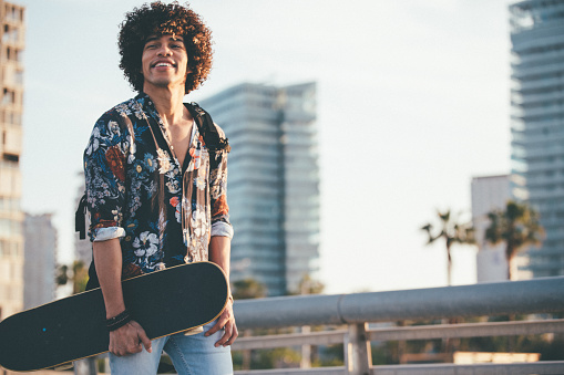 One man, curly haired young man, holding skateboard on the street on a sunny day.