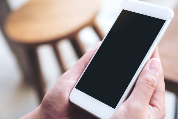istock Mockup image of hands holding white mobile phone with blank black desktop screen in cafe 973158710