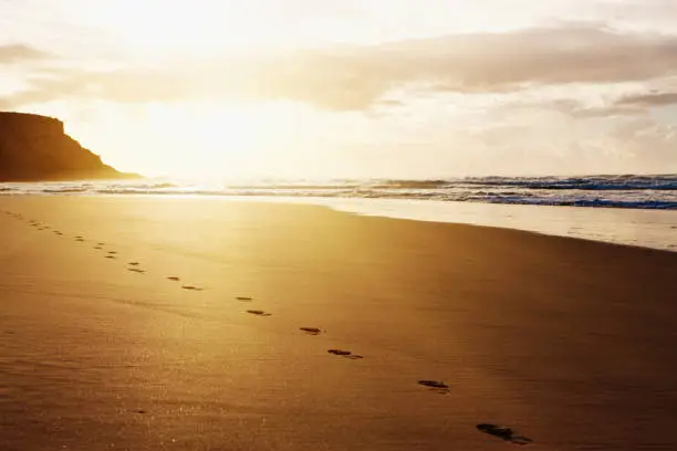 Photo of Sunrise or sunset highlights footprints of a lone beach stroller