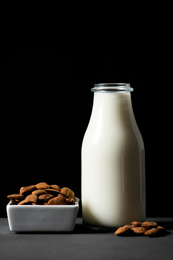 closeup of a white ceramic bowl with almonds and a glass bottle with almond milk on a gray rustic wooden table, against a black background, with some blank space on top