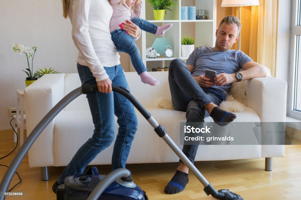 Woman with small child doing housekeeping while man sitting in couch Woman with small child in her hands doing housekeeping while man sitting in couch and relaxing. Women Stock Photo