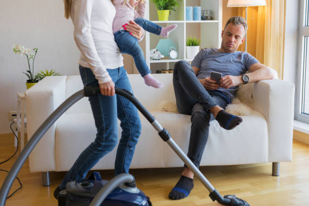 Woman with small child doing housekeeping while man sitting in couch Woman with small child in her hands doing housekeeping while man sitting in couch and relaxing. chores stock pictures, royalty-free photos & images