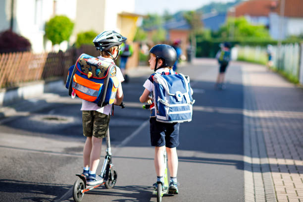 zwei school kid jungen in schutzhelm mit roller in der stadt mit rucksack an sonnigen tag reiten. glückliche kinder in bunten kleidern, radfahren am weg zur schule. - people child twin smiling stock-fotos und bilder