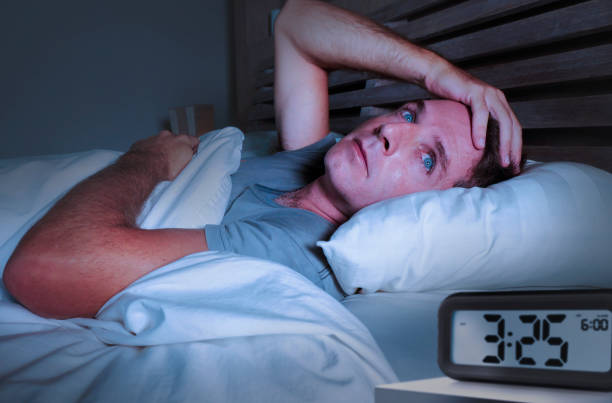 istock worried sad young attractive man awake lying sleepless on bed with eyes wide opened suffering insomnia sleeping disorder depressed and thoughtful with alarm clock pointing late night hour 973262392