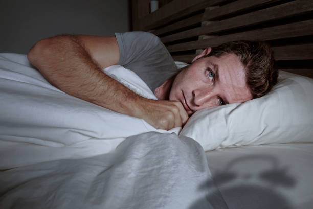 istock restless worried young attractive man awake at night lying on bed sleepless having eyes opened depressed suffering insomnia sleeping disorder with classic alarm clock shadow in problem concept 973262662