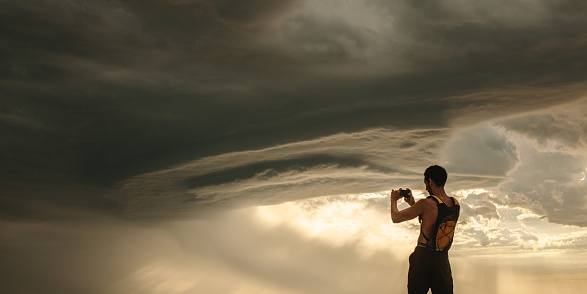 Rear view of athlete taking pictures of dramatic sky with his mobile phone. Fit man capturing cloudy sky pictures in his mobile phone while workout break.