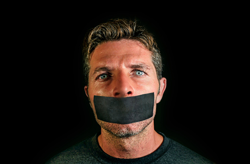 young man with mouth and lips sealed covered with adhesive tape in censorship coerced freedom of speech and forced silence and secrecy concept isolated on dark black background