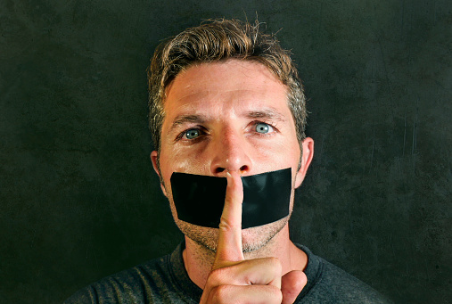 young man with mouth and lips sealed covered with adhesive tape in censorship coerced freedom of speech and forced silence and secrecy concept isolated on dark grunge background
