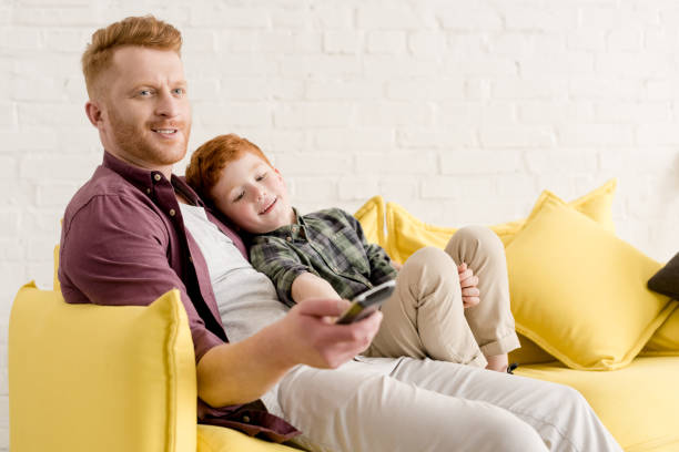 istock happy father and son sitting together on sofa and using remote controller while watching tv at home 973280740