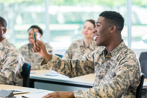 Confident African American male cadet raises hand to ask or answer question while in military academy class. Students are in the background.